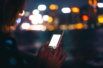 Crop young woman touching smartphone with copy space screen near blurred dark river and city at night