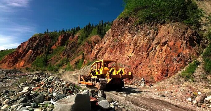 Large powerful yellow mining bulldozer operating and driving on brown dirt unpaved rocky remote rural road by red bedrock hills and green Yukon landscape, Canada, aerial tracking