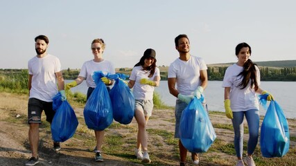 Multiracial volunteers charismatic young group beside of a big lake on the beach after they finish to cleaning up the rubbish happy take the plastic blue bags and start walking in front of the camera - Powered by Adobe