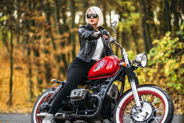 Obraz na płótnie Canvas Pretty blonde biker girl in sunglasses with red motorcycle on the road in the forest