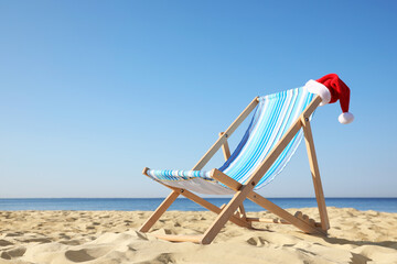 Deck chair with Santa Claus hat on sandy beach. Christmas vacation