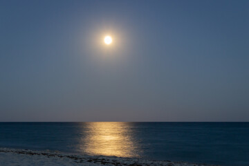 Full moon with moonlight on calm sea surface, long exposure. Island beach in the night. Moon path on sea. Twilight on seacoast. Scenic night sky over beach. Tranquility of summer night.