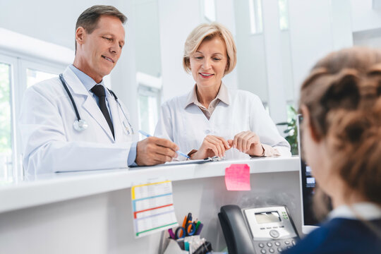 Two Doctors Colleagues Standing At Reception Desk Talking And Sharing Information With Receptionist In Modern Clinic