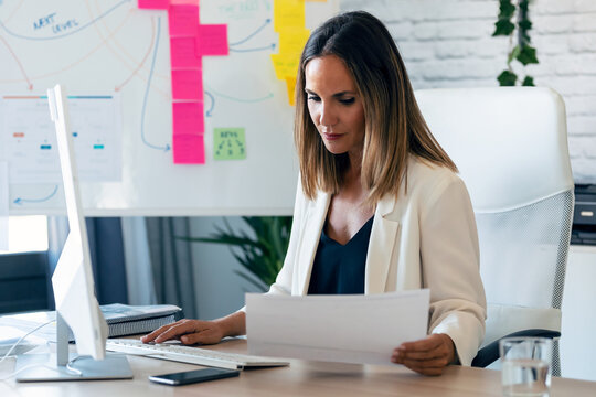 Confident business woman working with computer while reading some notes in the office.