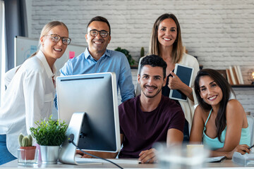 Successful business team standing around computer for presentation of work while looking at camera in the coworking space.