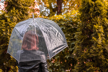 Autumn. Lonely redhead girl under a transparent umbrella with rain drops walking in a park, garden. Rainy day landscape