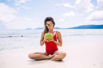 Fototapeta na wymiar Caucasian woman in red swimsuit drinking coconut beverage during sunbathing on tropical Hawaii beach, young female in sunglasses enjoying healthy vegan food during summer vacations on Seychelles