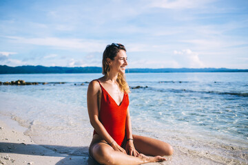 Side view of cheerful female resting at beach coastline and smiling during touristic journey on Bahamas, happy Caucasian woman enjoying Maldives travel vacations sitting at seashore with white sand