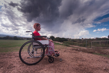 wide angle disabled girl sitting in a wheelchair in the middle of a green field in the countryside with a sky clouds
