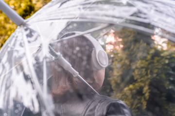 Autumn. Lonely sad woman in a headphones walking in a park, garden. View through wet  transparent umbrella with rain drops. Rainy day landscape.