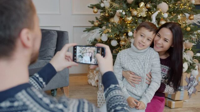 Father is taking picture of happy mother and son near Christmas tree using smartphone camera in beautiful decorated apartment. People and holidays concept.