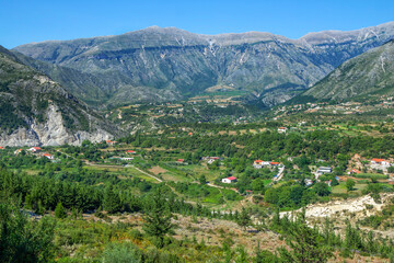 Fototapeta na wymiar View of valley and small village in Albanian mountains. Summer landscape with green trees, road on a hill and clouds on the sky. 