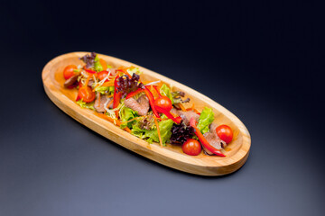 Beef Thai salad with herb mix and cherry tomatoes served in wooden plate on dark background. Side view of fresh salad with veal pieces, carrot and bell pepper slices. Restaurant menu dish isolation
