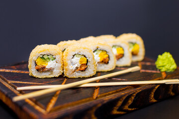 Close up of hot tempura roll with unagi eel, cucumber, Japanese omelette (tamago) and cream cheese served on wooden board. Pan Asian dish served with wasabi sauce and chopsticks on dark background
