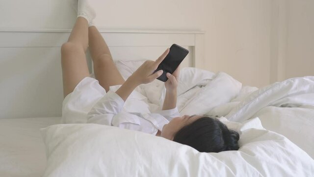 Asian beautiful woman holding smartphone shopping online, social media and fashion news at home. Teenages girl lying on bed and happy with news online in internet while being quarantine at home.