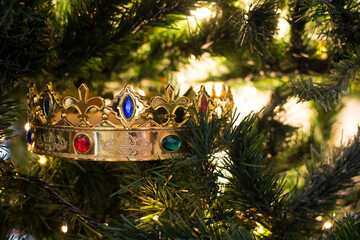 A golden wizard king crown with colored gems, among the branches of a Christmas tree illuminated by its Christmas lights.