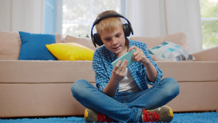 Boy kid in headphones playing game on smartphone comfortably sitting on floor