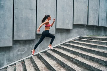 Tuinposter Full length of young woman in sports clothing jogging while exercising on the steps outdoors © gstockstudio