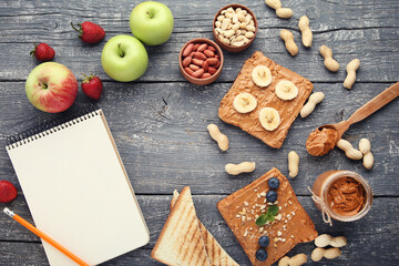 Bread with peanut butter, fruits, nuts and blank sheet of paper on wooden table