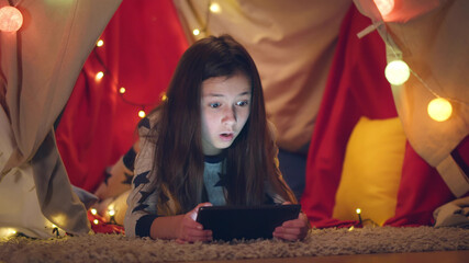 Smiling girl kid lying in cozy wigwam and using digital tablet