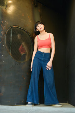 Outdoors Fashion. Beautiful And Young Model Woman With Short Hair And With Slim Perfect Body, In Pink Sleeveless Shirt And Wide Leg Blue Jeans Posing On Vintage Background.