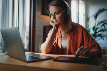 Home Office Concept, Young female freelancer working remotely from home wearing headphones using...