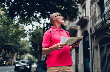 Mature male tourist in casual clothing using location map for search direction route during city sightseeing, aged man in classic spectacles with paper orientation in hands looking around