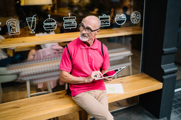 Pensive male blogger 60 years old holding modern touch pad and thoughtful looking away, contemplative man in optical spectacles with digital tablet feeling pondering on idea for web publication