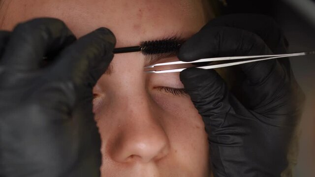 Professional styling, correction and lamination procedures of girl eyebrows in beauty salon. Closeup view of client face and stylist's hands working in gloves. Master removes hair using tweezers.