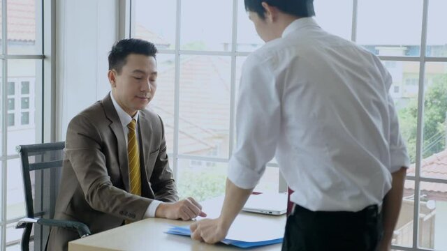 Indoor, Asian business concept. Male Subordinate in white shirt hands over portfolio and talks to his boss. Then Boss in professional suit receives the folder and look at the file, nods and smiles.