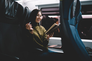 Smiling asian hipster girl reading book during trip in comfortable train sitting in seat holding literature, cheerful female blogger reading article for publication satisfied with trip 