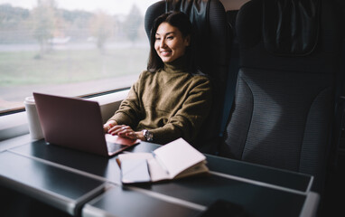 Cheerful asian female passenger of train typing on laptop computer working remotely during trip...
