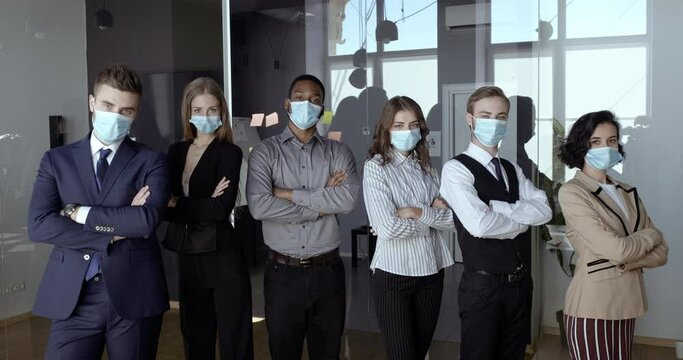 Motivated multidimensional business team stands together in conference room, confident colleagues fold arms over chests, office workers wear protective medical masks preventing virus, safety concept