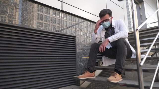 Caucasian young doctor man sits down on the stairs near the clinic building, tired and unhappy rubbing his nose and eyes, feeling tired and headache. Health care worker stress and frustration concept.