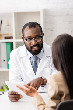 african american doctor in eyeglasses pointing with finger at digital tablet during conversation with patient