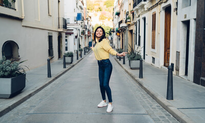 Fototapeta na wymiar Full length portrait of funny tourist in headphones walking around touristic town dancing and smiling at camera, happy female generation Z in electronic equipment enjoying pastime vacations for travel
