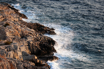Waves break on rocks on the seashore with white splashes. Blue foamy water on a stones, sea background