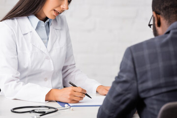 Fototapeta premium cropped view of doctor writing on clipboard near african american patient