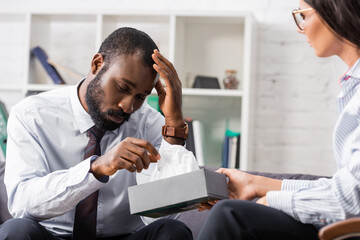 selective focus of frustrated african american man touching head while taking paper napkin from psychologist