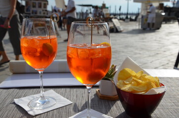 Two glasses of Aperol Spritz cocktails on a table in Venice Castello, Italy, salty crackers nearby, blurred waterfront background