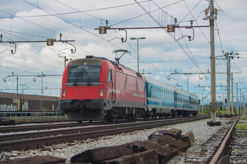 Fototapeta na wymiar Modern electrical train with red engine on track on a suburban station. Passenger train rushing towards the city in a suburban environment on a cloudy day.