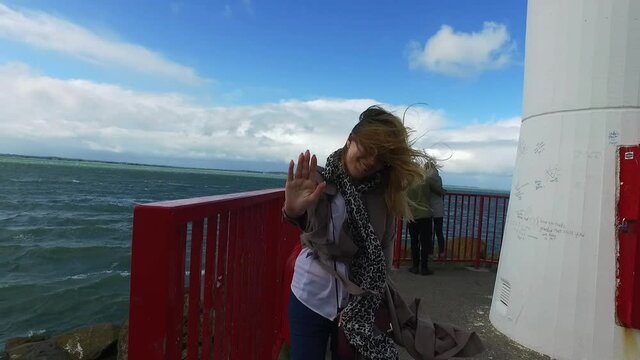 The girl walks on the pier to lighthouse in bad weather. A strong wind blows down a young woman. The fabric of the coat and scarf flutters. Storm at sea in Ireland near Dublin. High quality FullHD