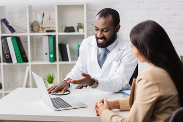 selective focus of bearded african american doctor pointing with hand at laptop near brunette woman