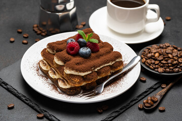 portion of Classic tiramisu dessert with raspberries and blueberries, cup of espresso and coffee maker on dark concrete background