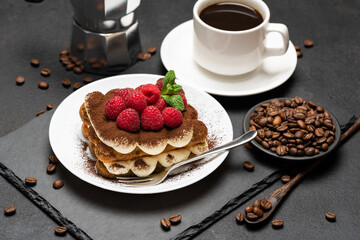 portion of Classic tiramisu dessert with raspberries, cup of espresso and coffee maker on dark concrete background