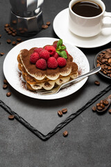 portion of Classic tiramisu dessert with raspberries, cup of espresso and coffee maker on dark concrete background