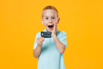 Excited happy little fun male kid boy 5-6 years old wearing stylish blue turquoise t-shirt polo hold in hand mockups of credit bank card isolated on yellow color wall background child studio portrait.