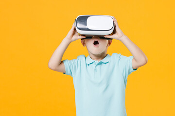 Smiling handsome little fun male kid boy 5-6 years old wearing stylish blue turquoise t-shirt polo watching in vr headset gadget on head isolated on yellow color wall background child studio portrait.