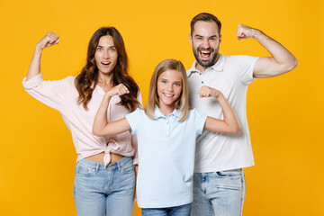 Strong funny young parents mom dad with child kid daughter teen girl in basic t-shirts showing biceps muscles isolated on yellow background studio portrait. Family day parenthood childhood concept.