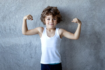 Little curly haired red boy in holding fists up and showing biceps on concrete grey background.